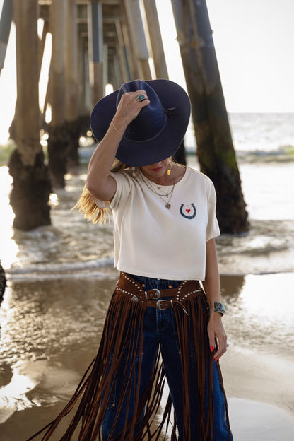 Person wearing a white t-shirt with a logo, blue jeans with fringe, and a dark hat on a beach.