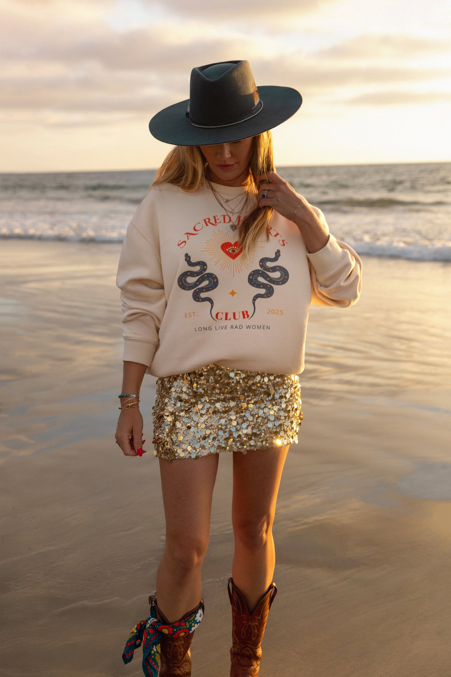 Woman on a beach wearing a beige sweatshirt with snake design, black hat, and sequin skirt.