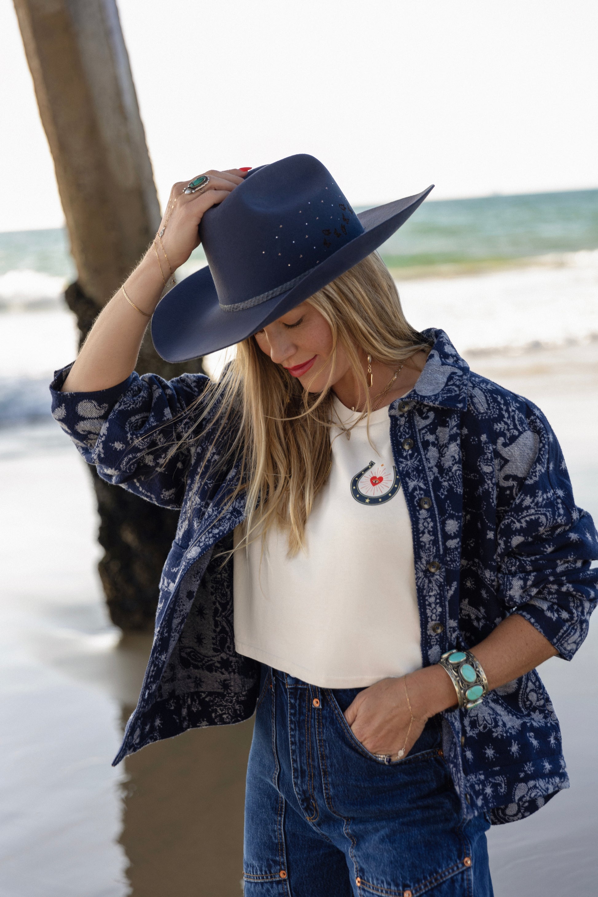 Person wearing a navy blue hat, patterned shirt, and jeans by the beach.
