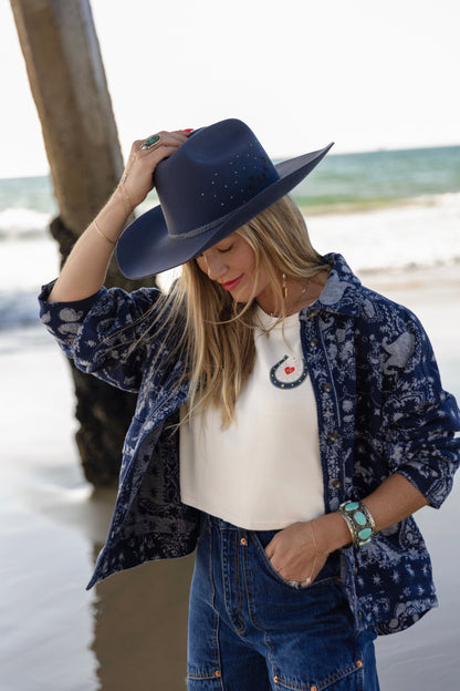 Person wearing a navy blue hat, patterned shirt, and jeans by the beach.