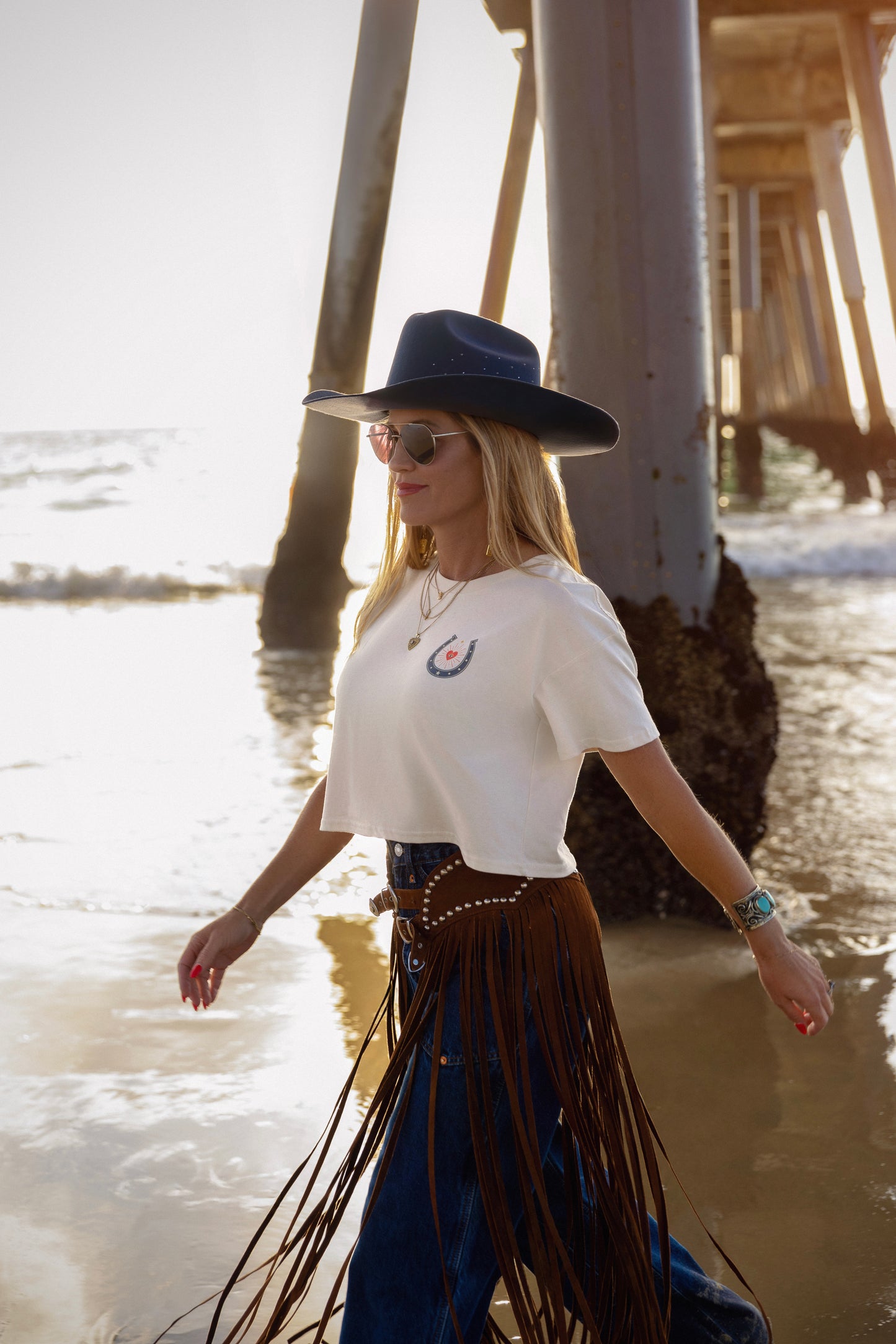 Woman in a white t-shirt and blue fringed skirt walking on a beach near a pier.