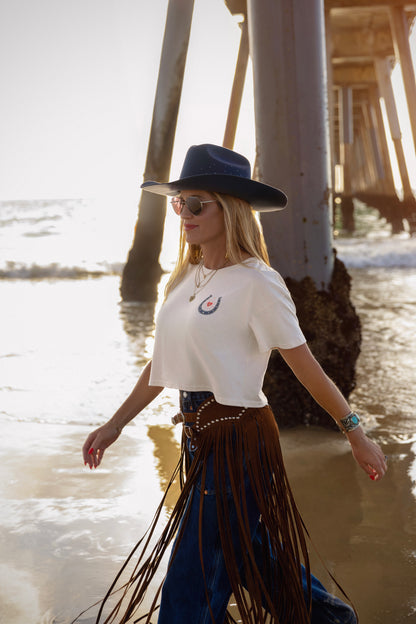 Woman in a white t-shirt and blue fringed skirt walking on a beach near a pier.