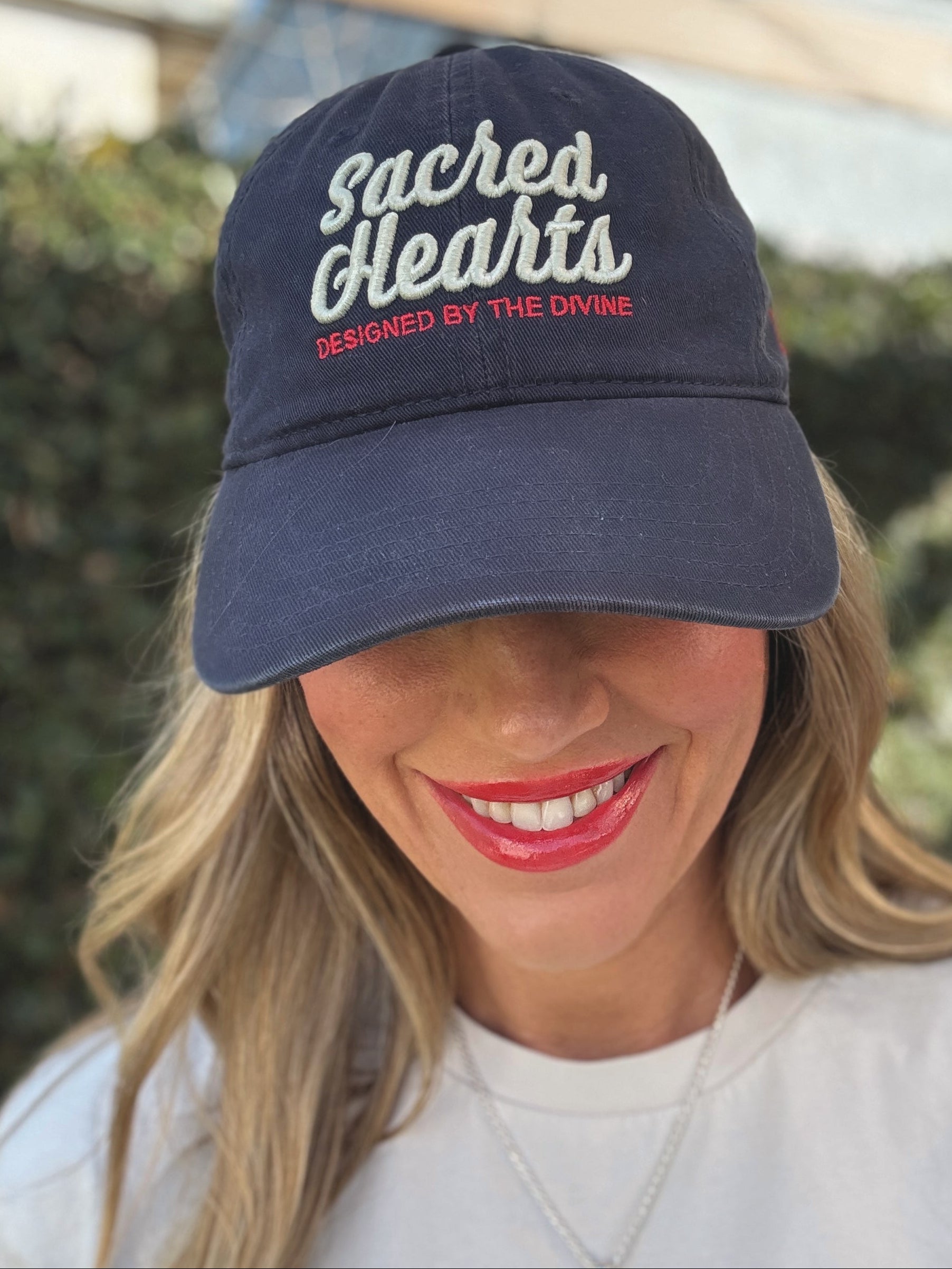 Woman wearing a navy cap with 'Sacred Hearts' text, smiling outdoors.